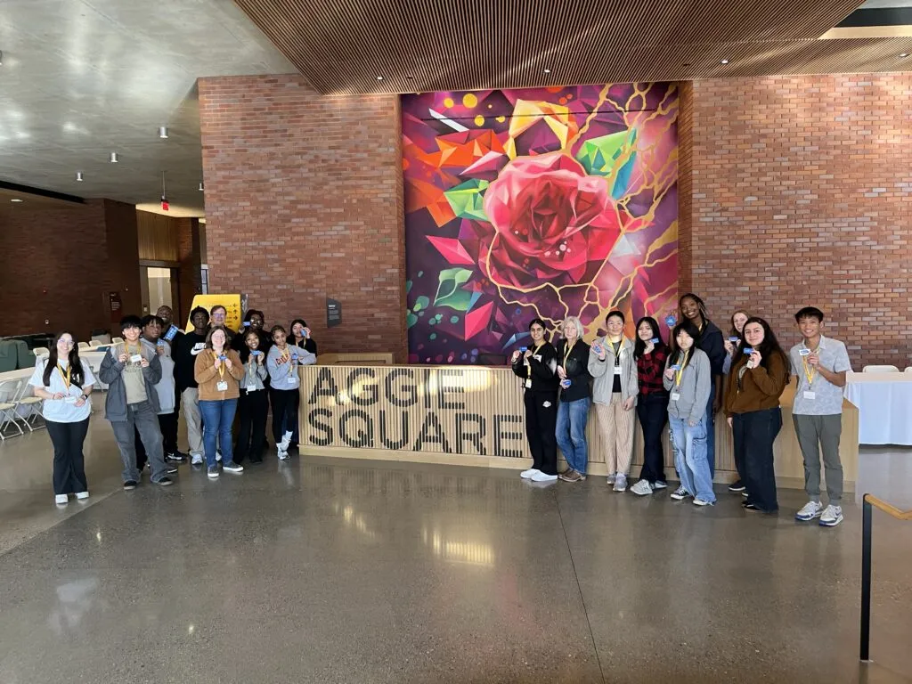 High school students hold up 3D-printed key chains as the pose in front of a colorful mural of a flower and a lobby desk with a sign that says 