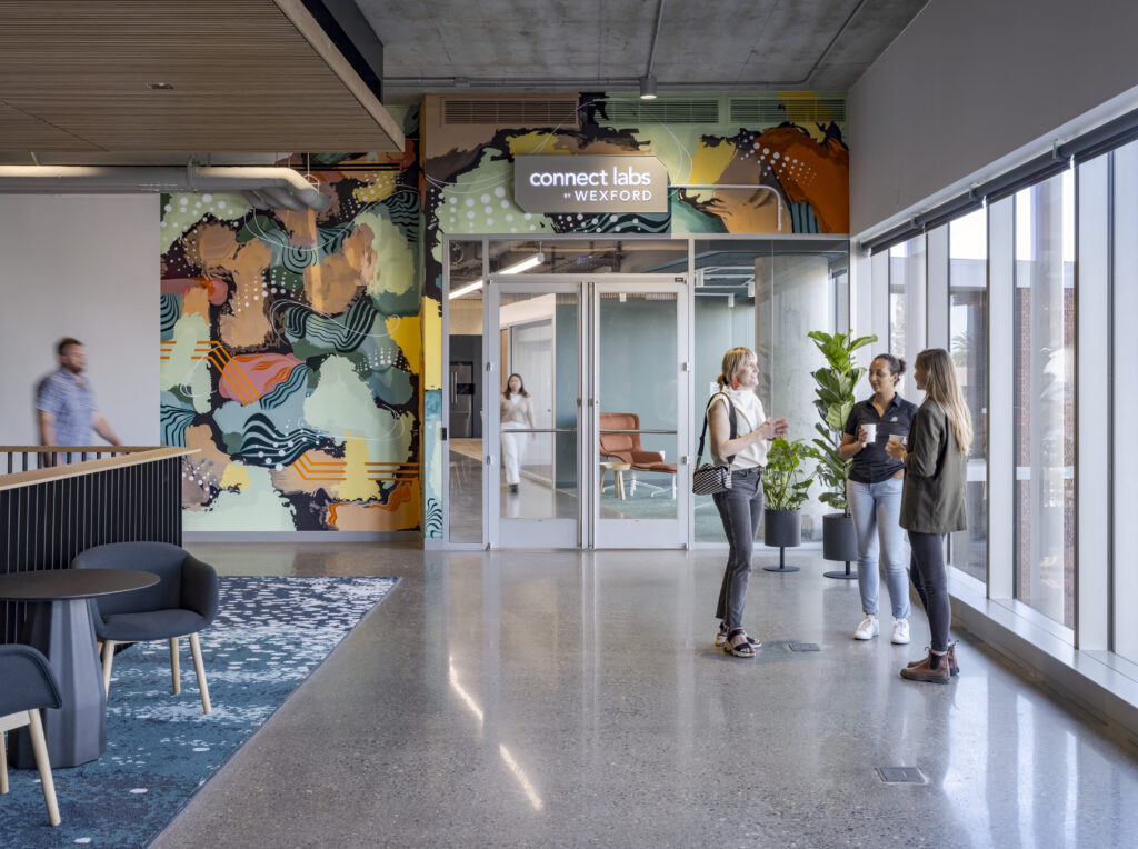 People mingle in a building lobby with a colorful mural in the background.