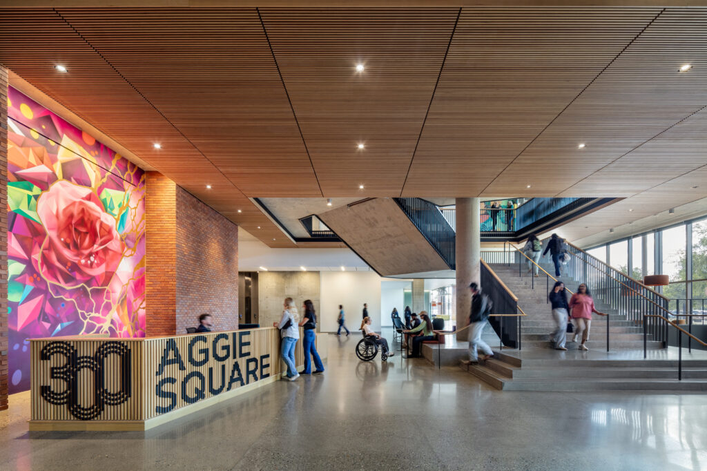 Modern lobby with colorful floral mural, stairs, glass wall and people by Aggie Square sign