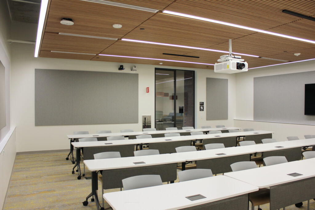 An empty classroom with rows of white tables and chairs and grey panels on the wall.